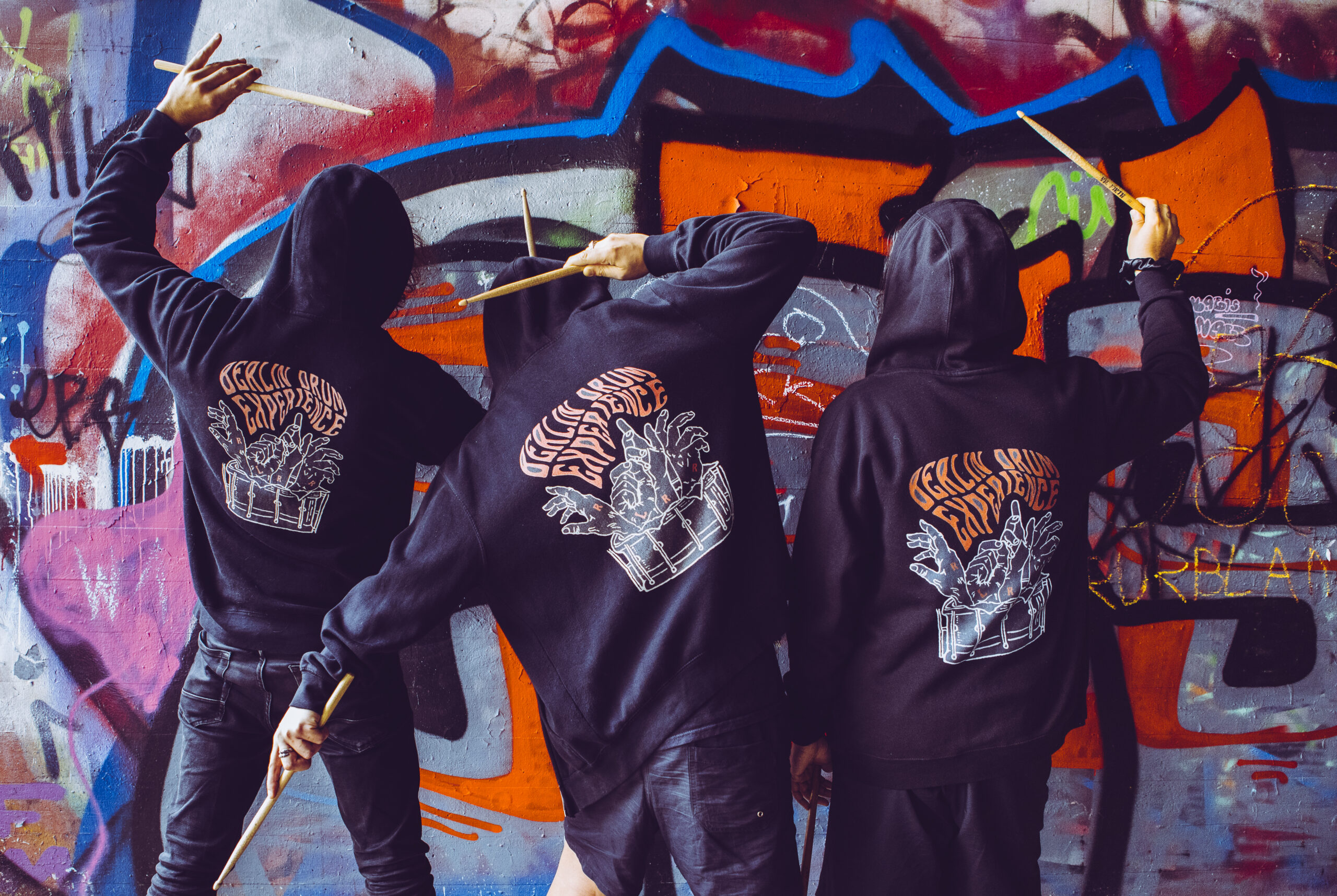Three BDE drummers raising their sticks beside a graffiti wall
